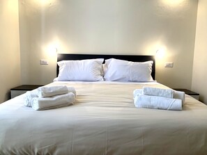 A neatly made bed with white linens and pillows, two bedside tables with lamps, and a plain wall. - Domo Achenza (Terralba)