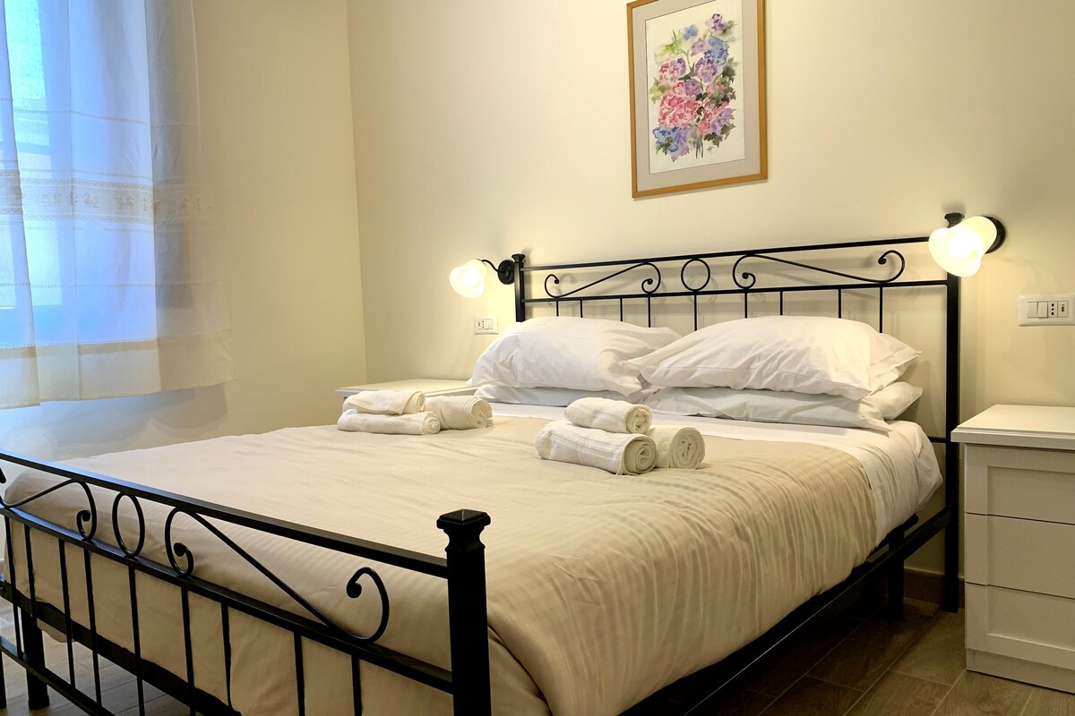 A neatly made bed with white linens and rolled towels, a black metal headboard, bedside lamps, and a framed floral artwork on the wall.