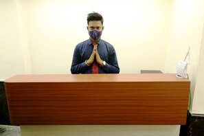 A person standing behind a wooden reception desk with a white device on the right. - The Lake Hotel (Kolkata)