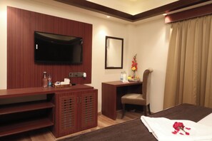 A hotel room with a flat-screen TV mounted on the wall, a wooden desk with a chair, a bed with a white bedsheet and a rose, and a large window with beige curtains. - The Lake Hotel (Kolkata)
