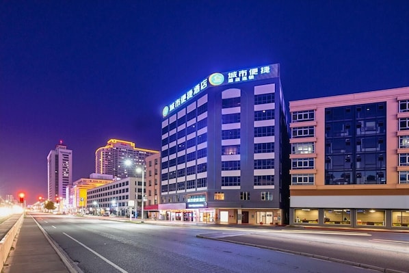 Un immeuble d’hôtel à plusieurs étages, doté d’une enseigne lumineuse, sous un ciel dégagé, et une rue où l’on aperçoit des traînées de lumière témoignant de la circulation.