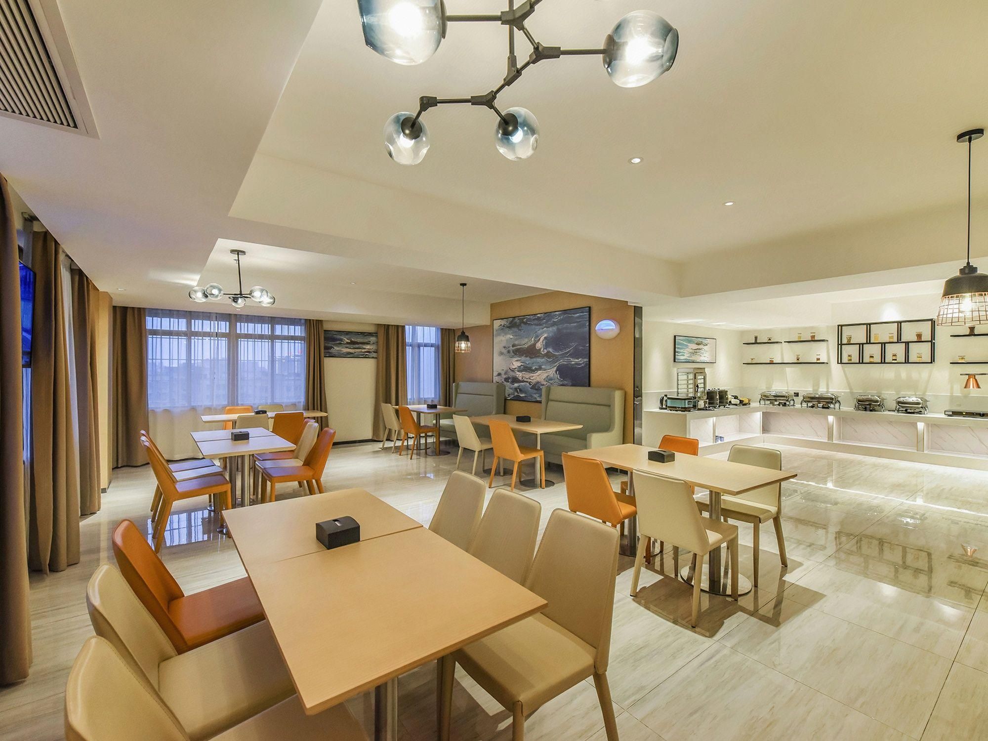 A hotel dining area with tables and chairs, a kitchen in the background, and a large painting on the wall.