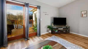A living room with a wooden floor, a TV on a stand, a sliding glass door leading to a balcony, and a small plant on the floor.