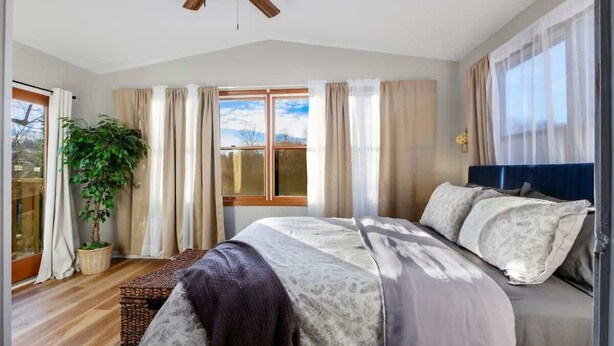 A bedroom with a large bed, a ceiling fan, a window with curtains, and a potted plant.