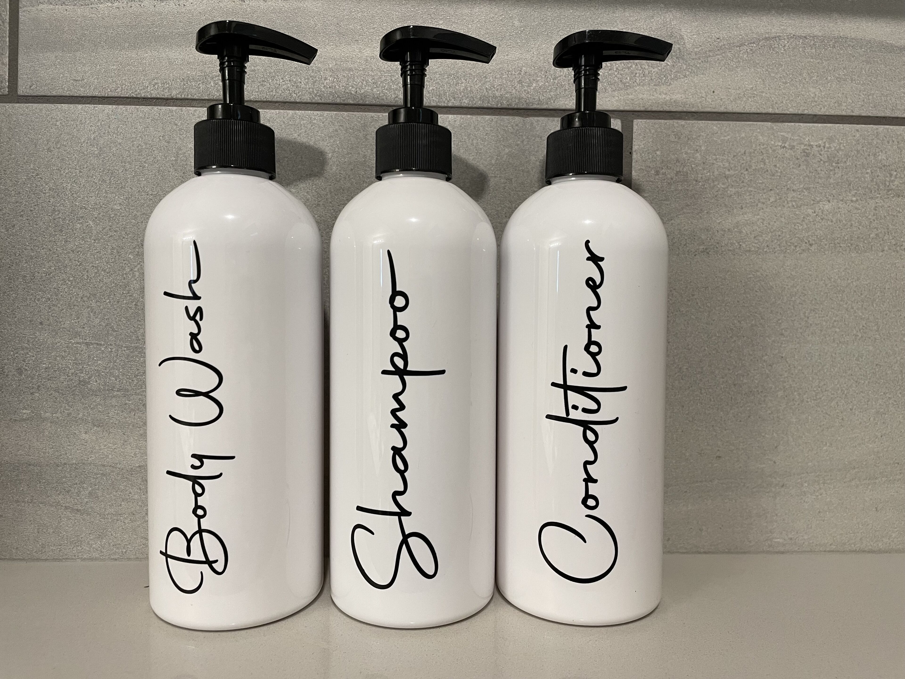 Kolme valkoista pulloa, joissa on mustat pumppupullot ja joihin on merkitty tekstit ”vartalopesu”, ”shampoo” ja ”hoitoaine”.