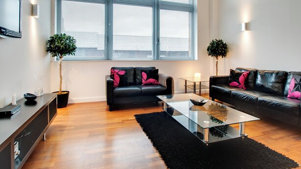 A modern living room with two black leather sofas, a glass coffee table, and a large window with blinds. - Crown Apartments 3A - UK3380 (Newcastle upon Tyne)