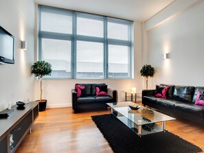 Un salon moderne avec deux canapés en cuir noir, une table basse en verre et une grande fenêtre équipée de stores.