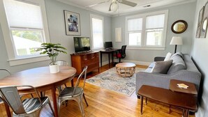 Uma sala de estar com uma mesa de jantar, cadeiras, um sofá cinza, uma mesa de centro, uma televisão e um ventilador de teto.