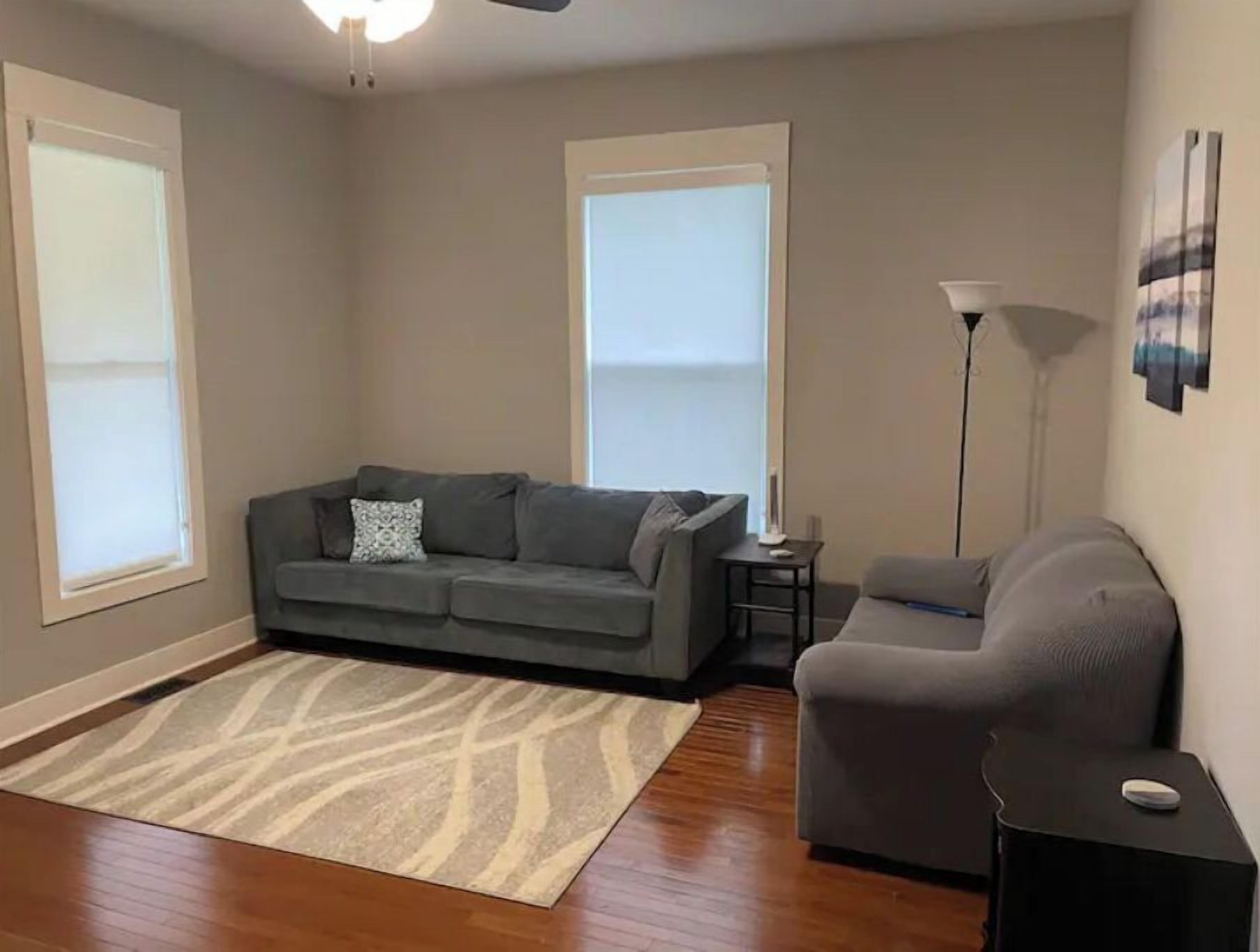 Un salon avec un canapé gris, un fauteuil gris, une petite table d’appoint, un lampadaire et un tapis à motifs.