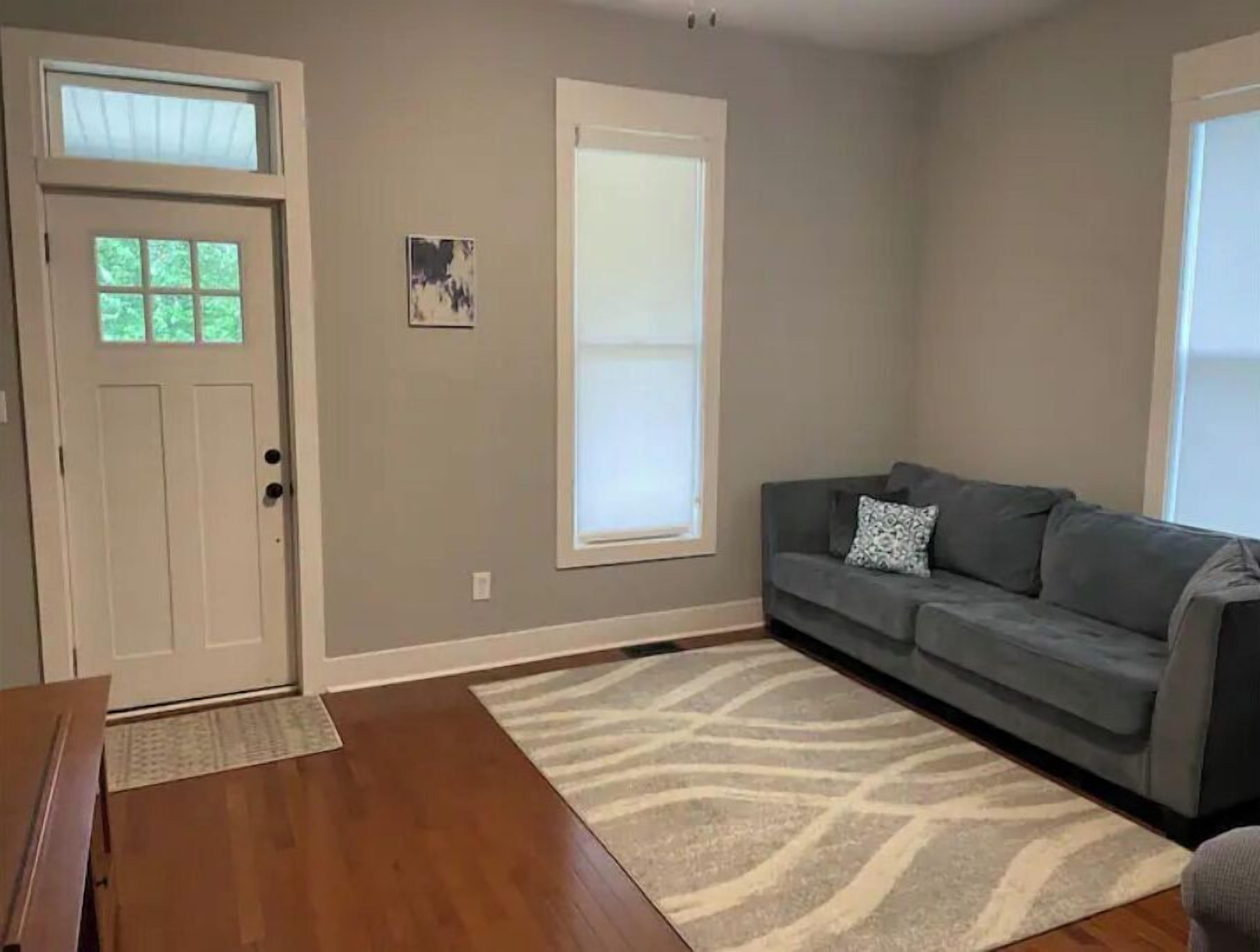 Una sala de estar con un sofá gris, un tapete estampado, piso de madera, una puerta blanca y una ventana con persianas.