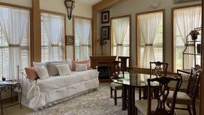 A cozy living room with a sofa, dining table, and chairs. There are large windows with curtains, a ceiling fan, and a fireplace.