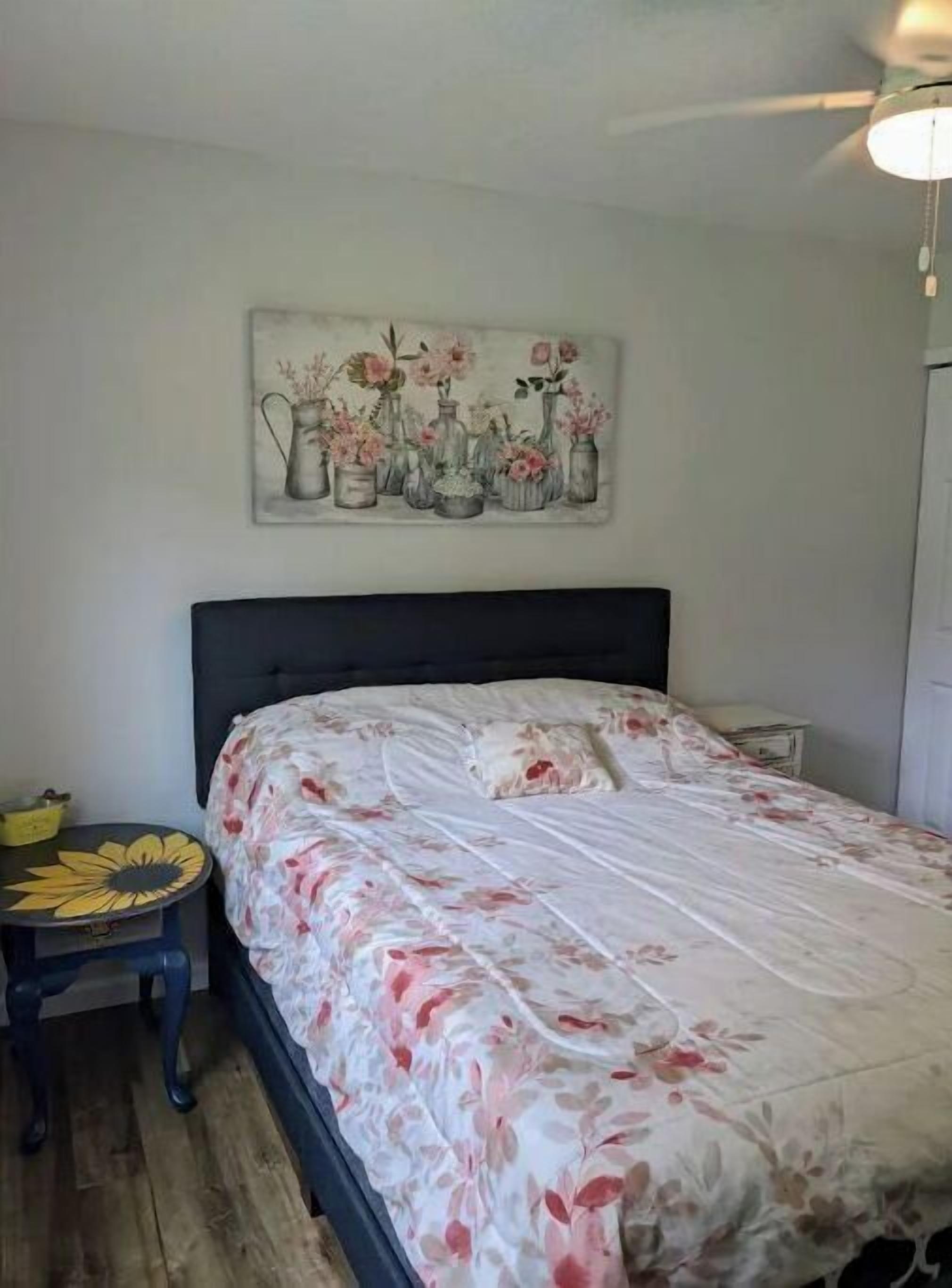 Et soveværelse med en seng, et natbord, et lille bord med en solsikke-dekoration og et blomster-maleri på væggen.