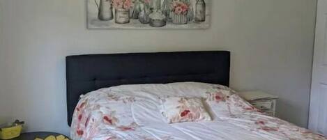 Et soveværelse med en seng, et natbord, et lille bord med en solsikke-dekoration og et blomster-maleri på væggen.