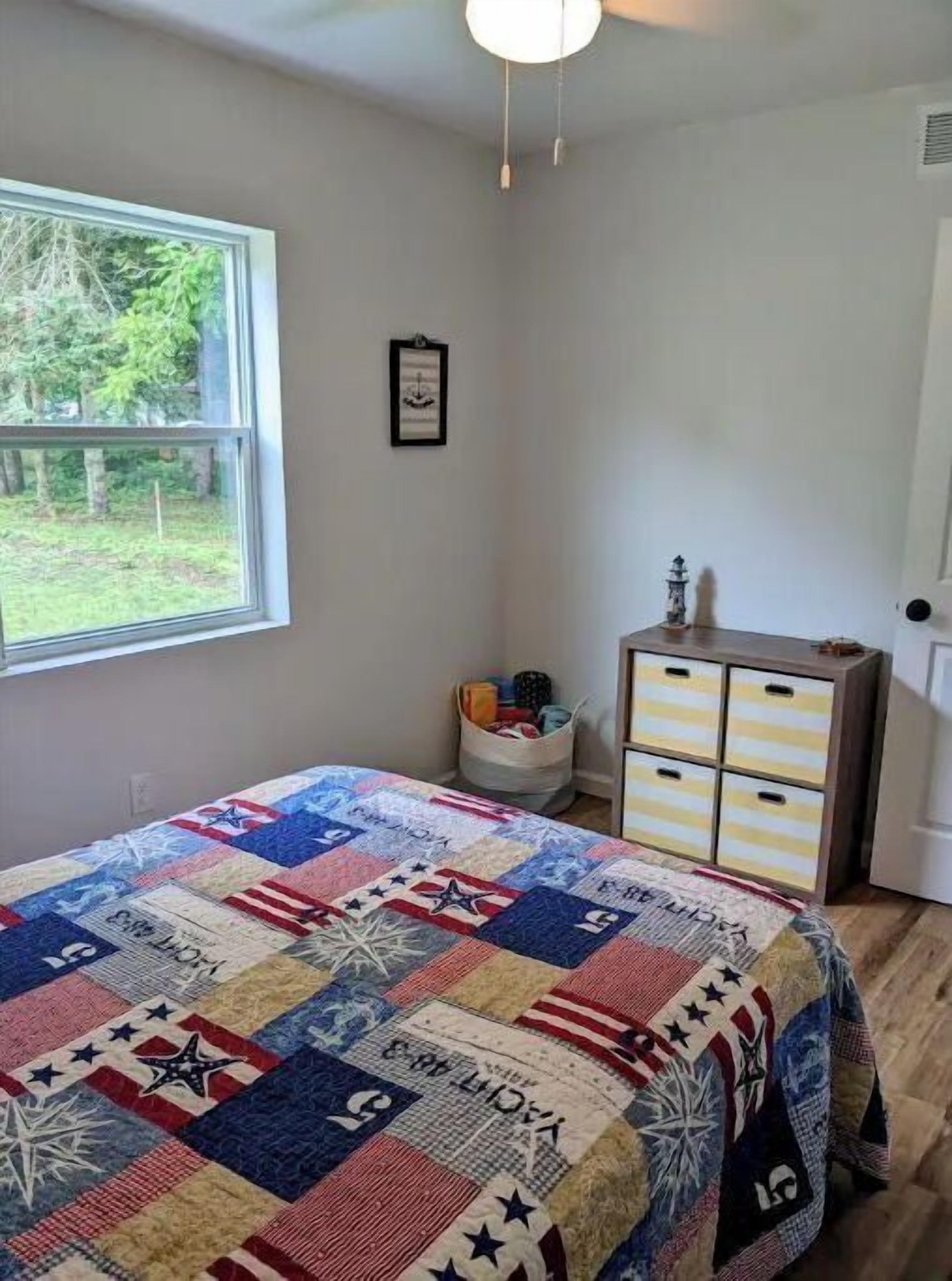 Ein Schlafzimmer mit großem Fenster, einem Bett mit Patchworkdecke, einem Schminktisch und einer kleinen Kommode.