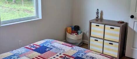 Ein Schlafzimmer mit großem Fenster, einem Bett mit Patchworkdecke, einem Schminktisch und einer kleinen Kommode.
