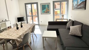 Un soggiorno moderno con un divano, un tavolino e un tavolo da pranzo con sedie. Sono presenti due finestre che offrono vista sugli edifici esterni.