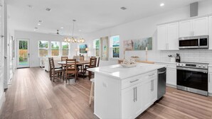 A modern kitchen with white cabinetry, a central island, and a dining area with wooden chairs. - Bliss: Gated! Pool! Free Golf Cart! Sleeps 20! (Miramar Beach)