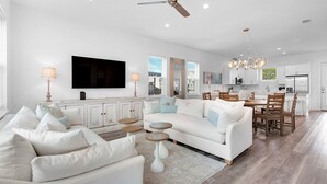 A spacious living room with a white sofa, a television, and a dining area with wooden chairs and a table. - Bliss: Gated! Pool! Free Golf Cart! Sleeps 20! (Miramar Beach)
