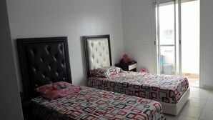 Un dormitorio con dos camas, cada una con una colcha estampada y un cabecero. Hay una mesita de noche con un espejo y un objeto rojo encima. Las paredes son blancas y hay un ventanal con vistas al exterior.