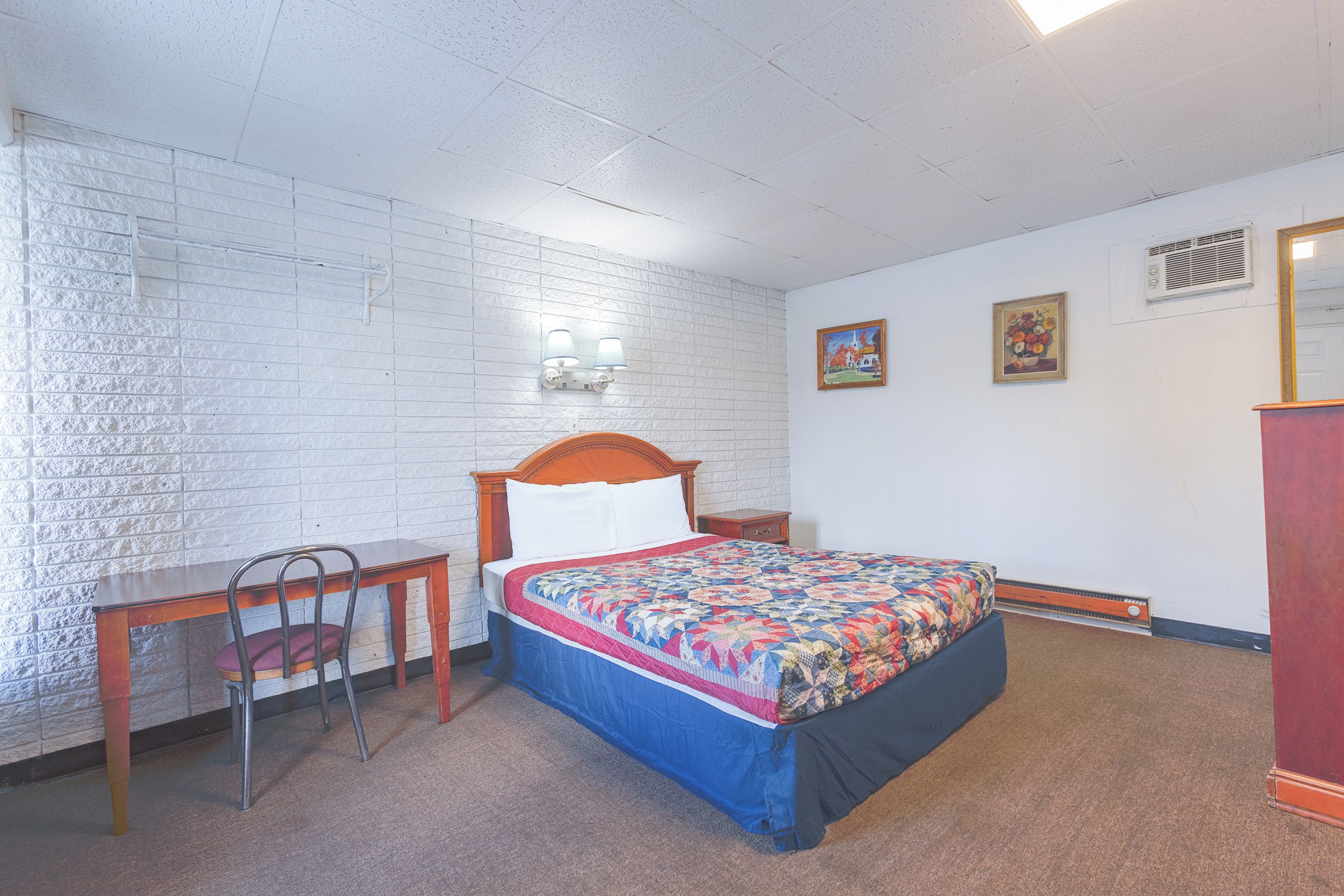 Une chambre à coucher avec un lit, un bureau et une chaise. Il y a deux lampes fixées au mur et deux tableaux encadrés accrochés au mur.