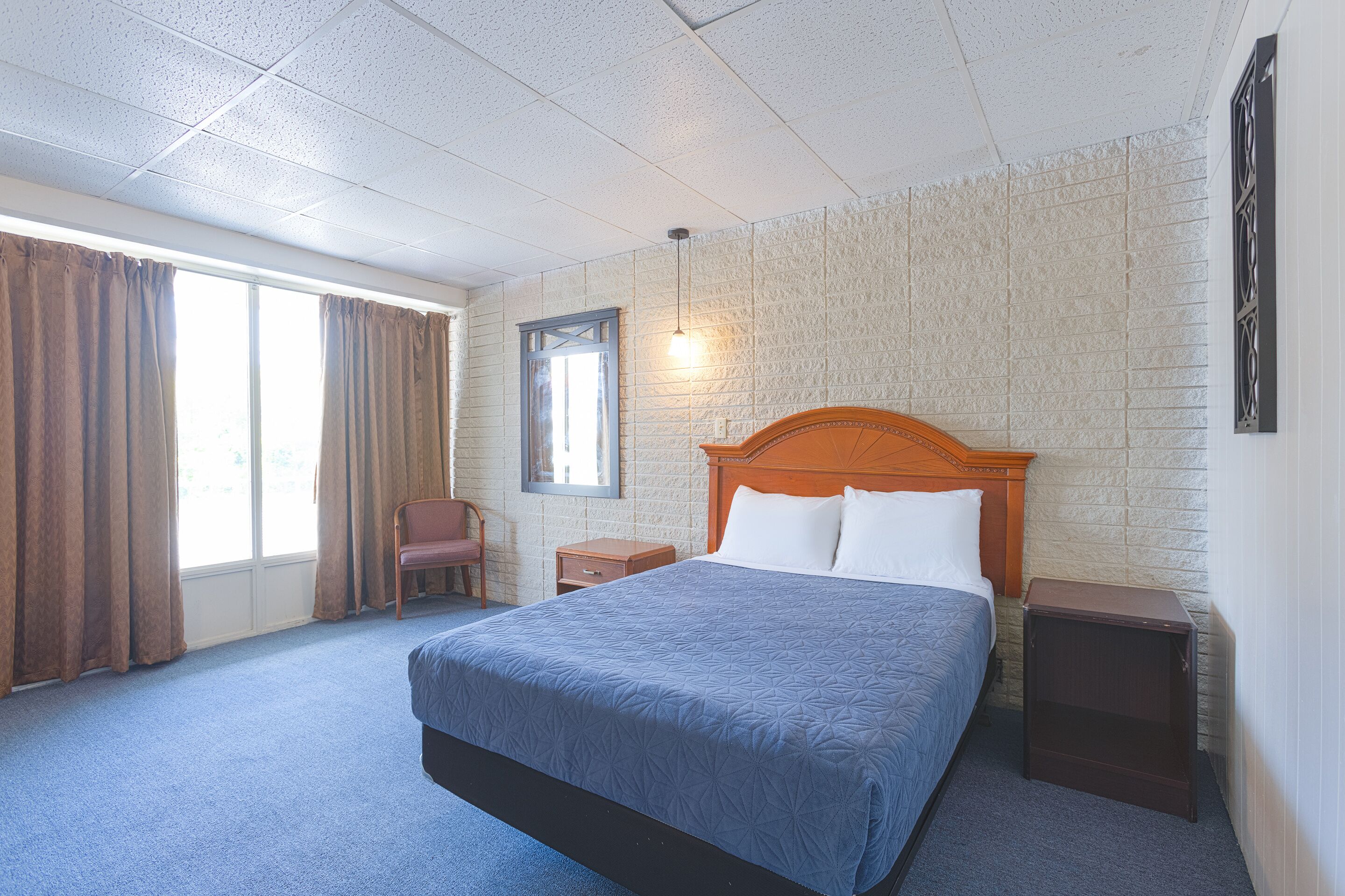 A hotel room with a large bed, a wooden headboard, a nightstand, a chair, a window with curtains, and a wall-mounted light fixture.