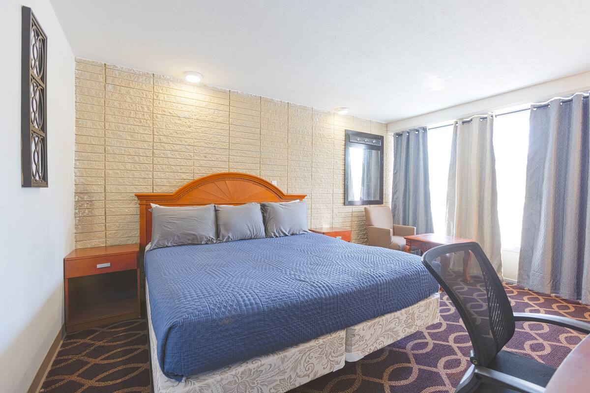 Una camera d'albergo con un letto dotato di poggiatesta in legno, un comodino, una sedia, un tappeto decorato e una parete con motivo a mattoni.