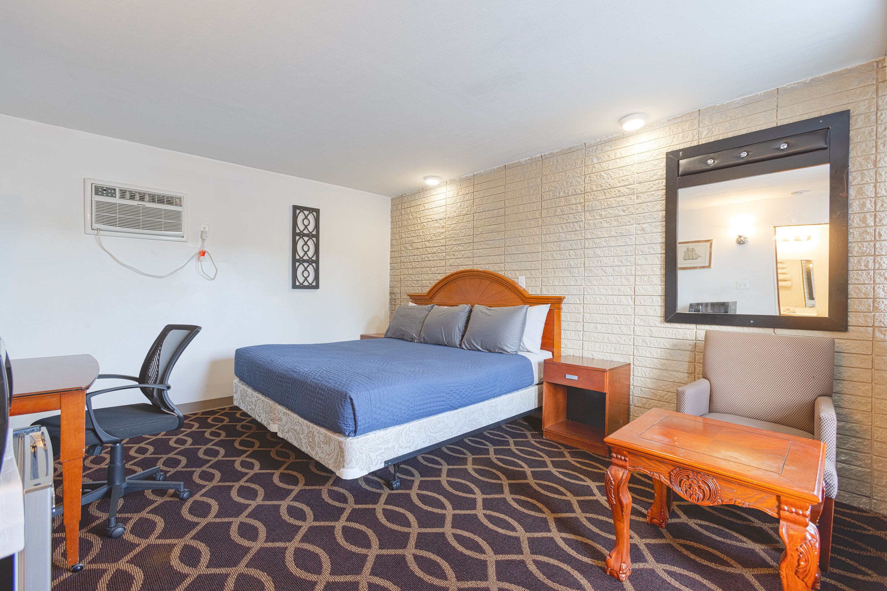 A hotel room with a bed, a desk, a chair, a nightstand, a mirror, and a patterned carpet.
