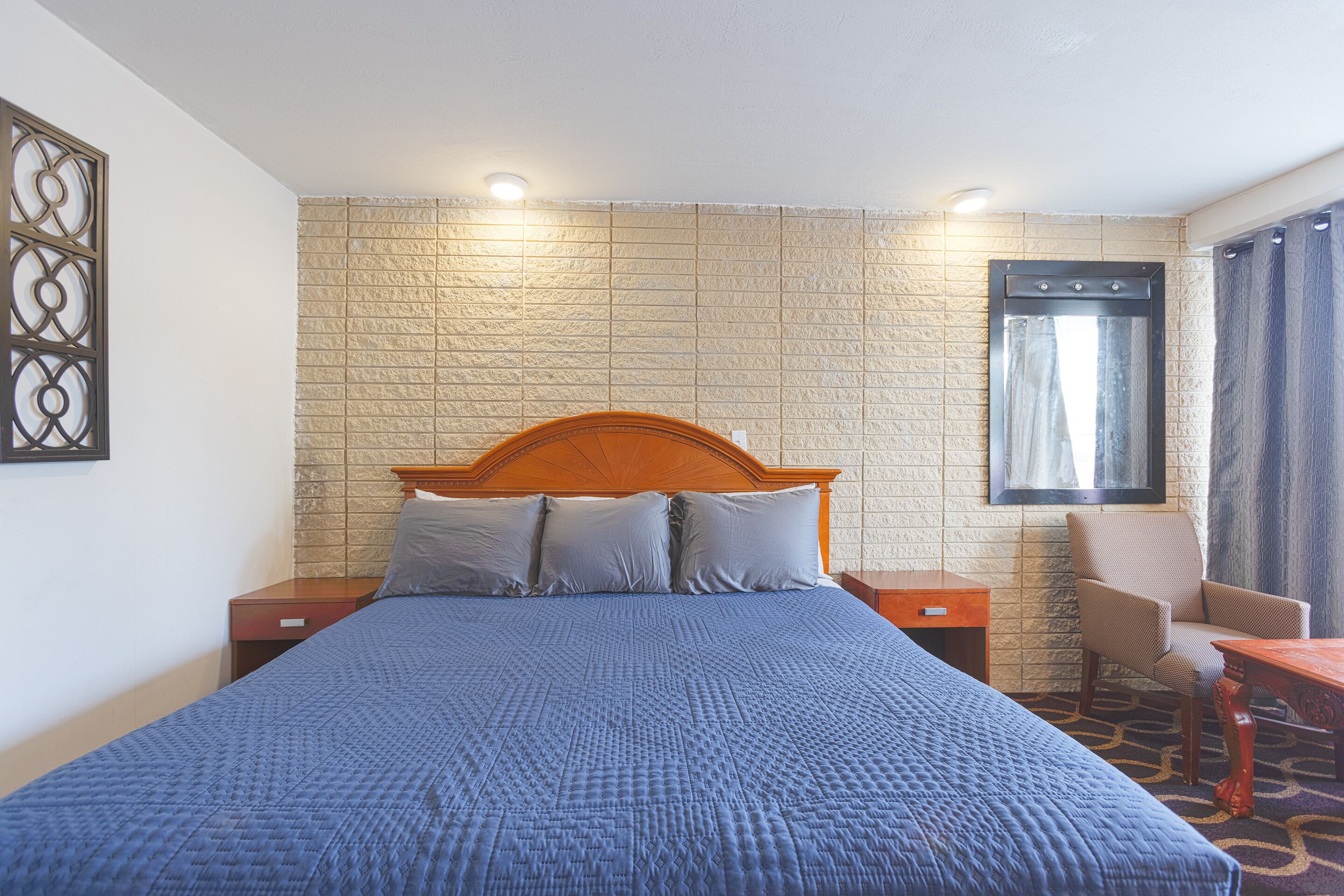 Ein Hotelzimmer mit einem grossen Bett, einem hölzernen Kopfteil, zwei Nachttischen, einem Sessel, einem kleinen Tisch und einem Fenster mit Vorhängen.