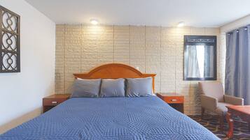 Ein Hotelzimmer mit einem grossen Bett, einem hölzernen Kopfteil, zwei Nachttischen, einem Sessel, einem kleinen Tisch und einem Fenster mit Vorhängen.