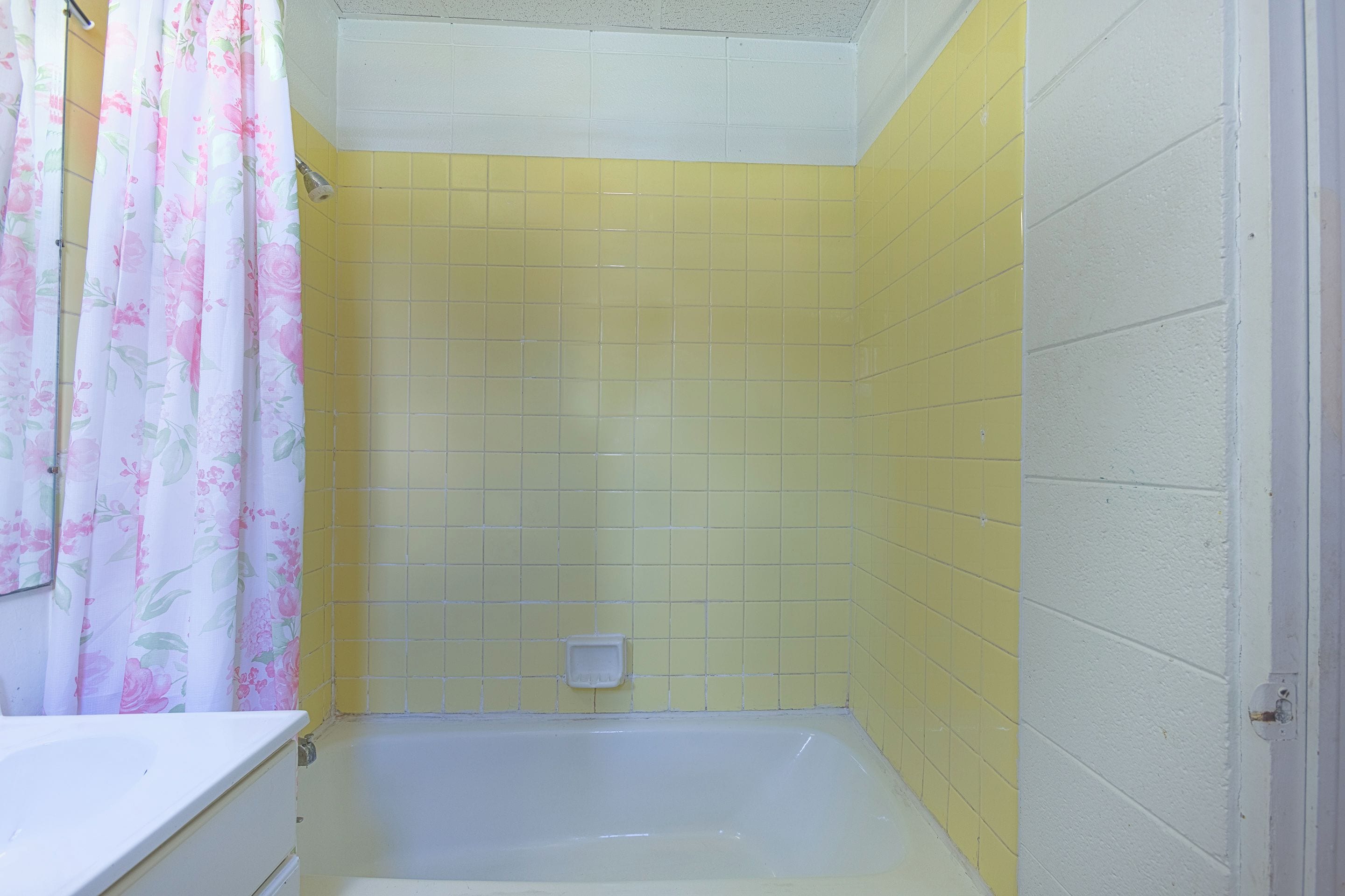Une salle de bain avec des murs recouverts de carreaux jaunes, une baignoire blanche et un rideau de douche à motifs floraux.