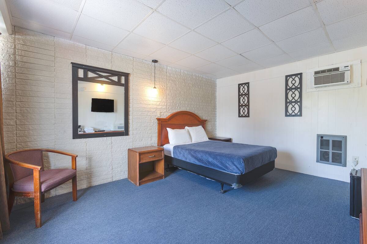 Una camera d'albergo con un letto, una sedia, un comodino, una TV a parete e una finestra con condizionatore.