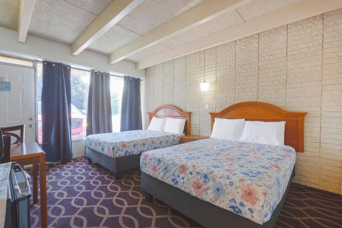 Camera d'albergo con due letti, una scrivania e una sedia. Si vede una finestra con tende, un tappeto a motivi e un muro in mattoni.