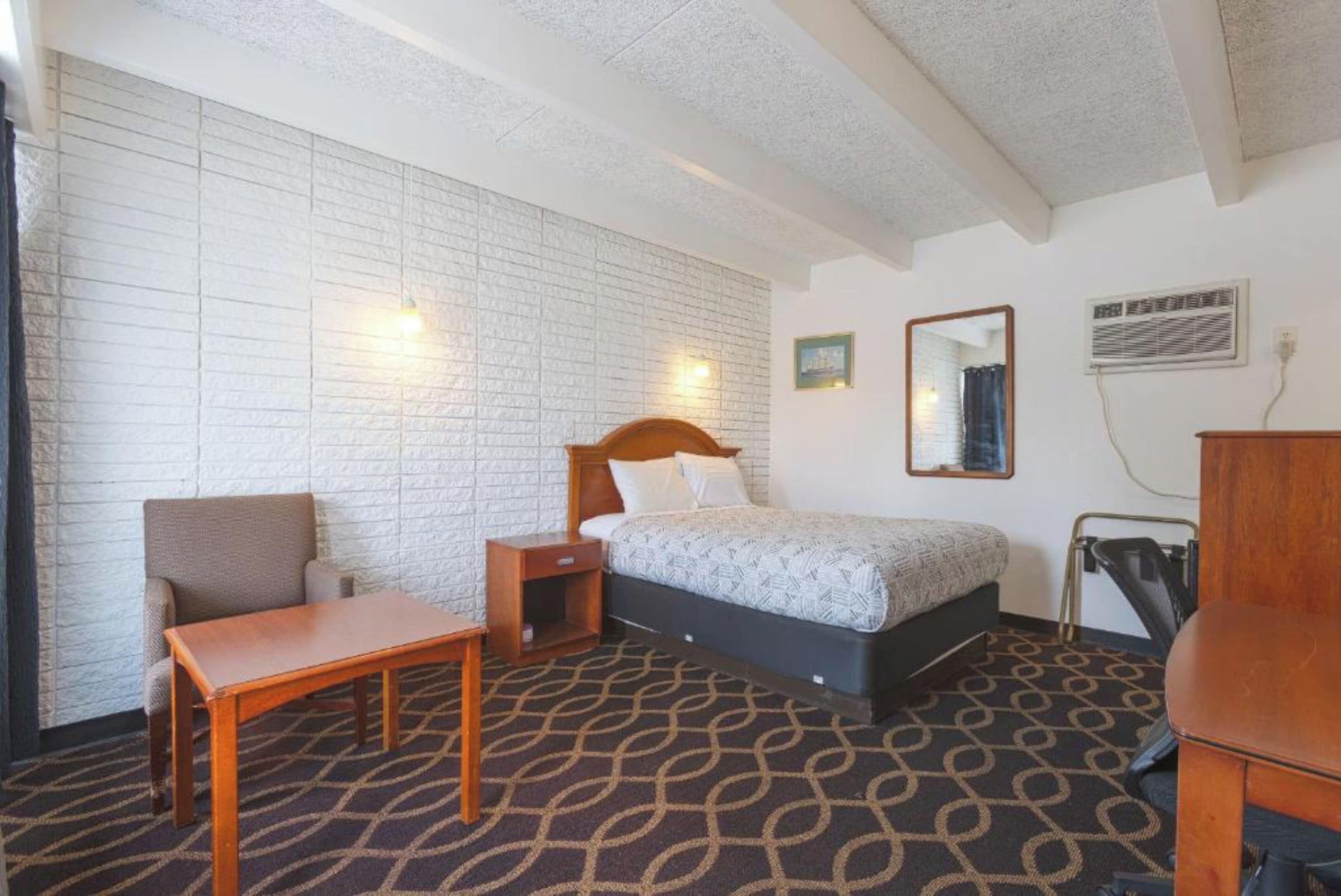 Una habitación de hotel con una cama, una mesita de noche de madera, una silla, una pequeña mesa y una alfombra estampada.