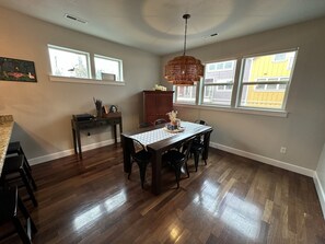 Un espace repas comprenant une table en bois, des chaises, un luminaire suspendu et une vue sur l’extérieur grâce à de grandes fenêtres.