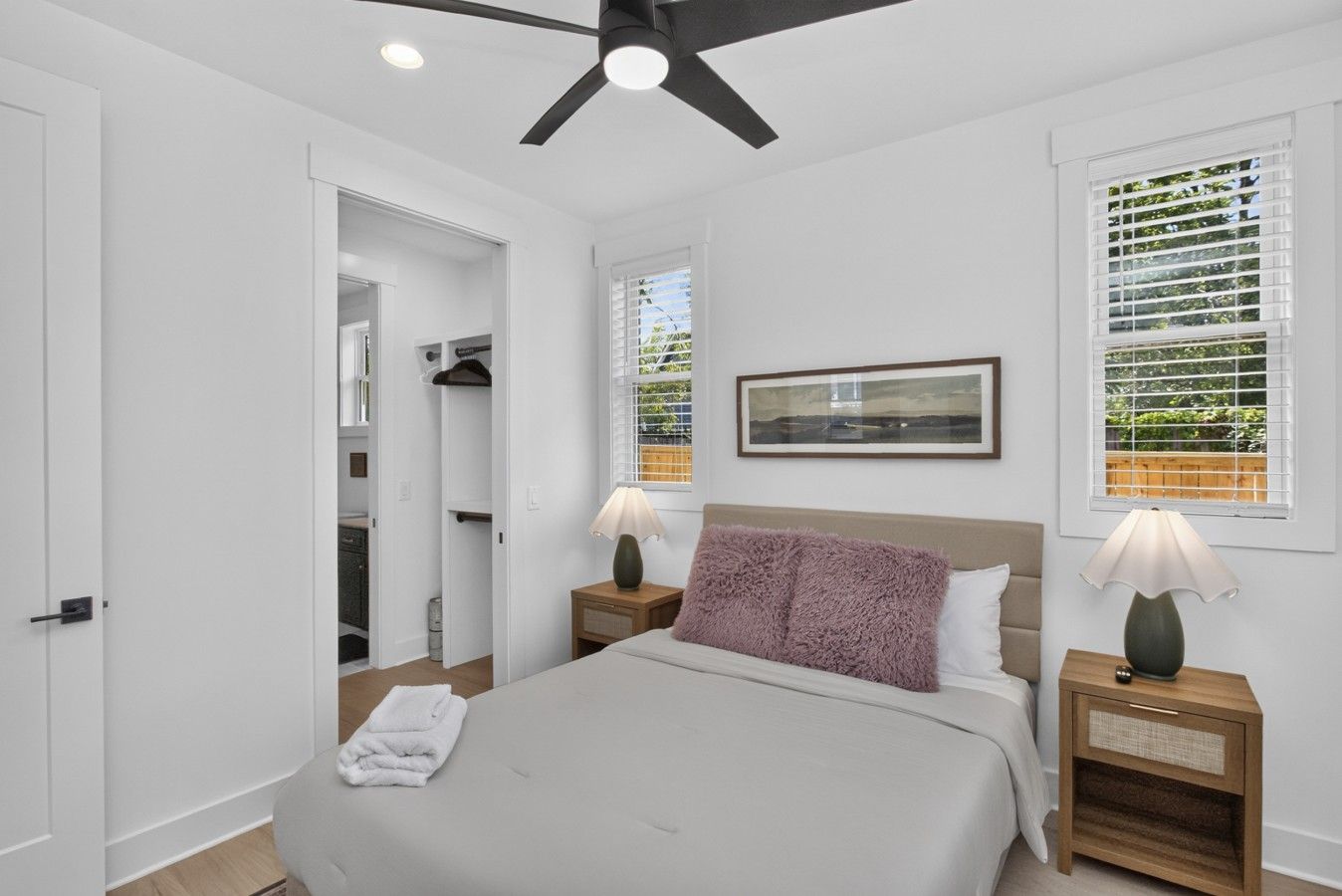 A bedroom with a bed, two bedside tables with lamps, a ceiling fan, a window with blinds, and a door leading to a closet.