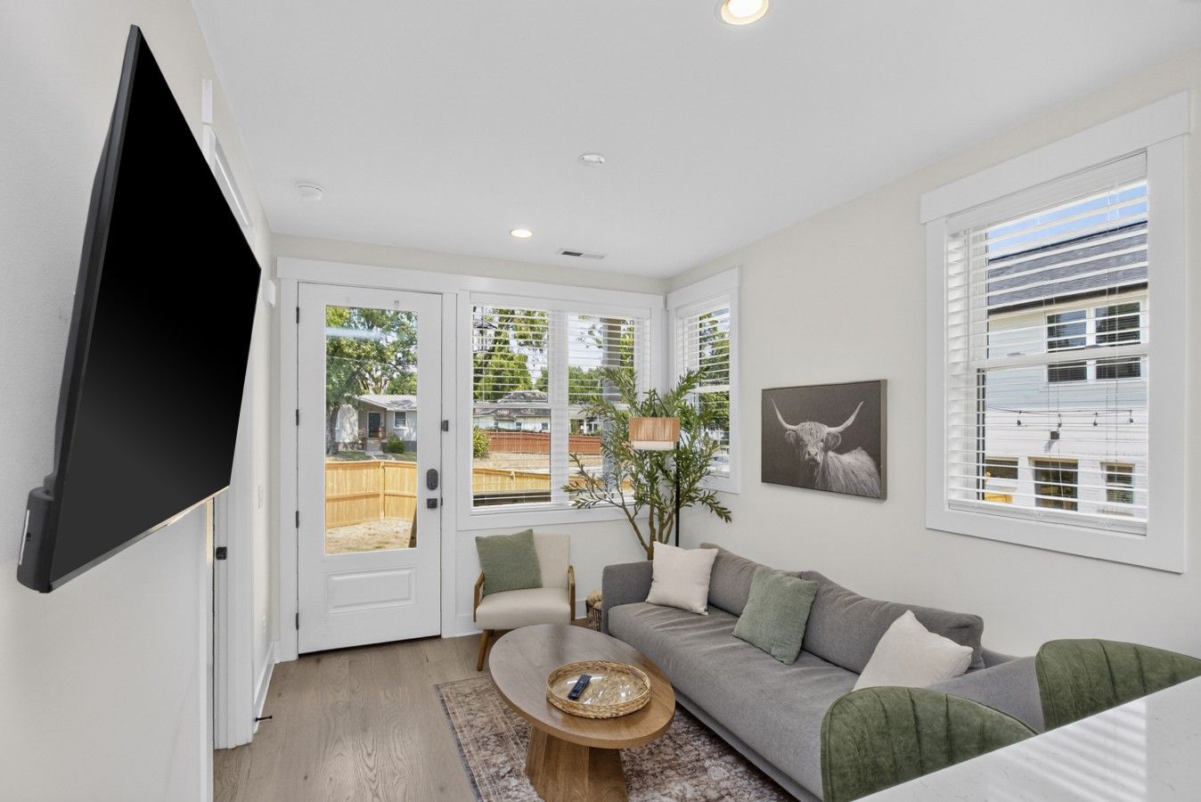 Ein modernes Wohnzimmer mit Fernseher, einem grauen Sofa, einem Couchtisch und einem großen Fenster mit Jalousien.