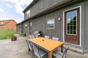 A wooden building with a patio area featuring a table and chairs, a grill, and a fire pit. - Sleeps 14 on a Duck River ranch: 4 en suite bedrooms, patio, fishing, horses (Normandy)