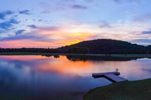 Ein ruhiger See mit einer anlegenden Mole, umgeben von Bäumen und einer Hügelkette im Hintergrund.