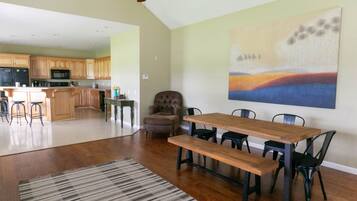 A spacious living area with a wooden dining table and chairs, a large abstract painting on the wall, and a kitchen in the background.