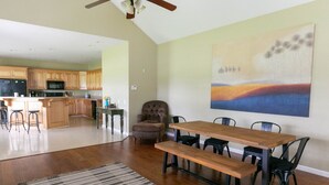 Uma sala de estar ampla com uma mesa de jantar de madeira e cadeiras, um quadro abstrato grande na parede e uma cozinha ao fundo.