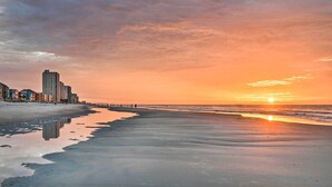 Une plage au coucher du soleil, avec des bâtiments en arrière-plan et une mer calme.