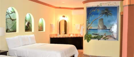 Une chambre d’hôtel avec deux lits, un ventilateur de plafond, une salle de bain avec un lavabo et un miroir, et une fresque représentant un paysage tropical.