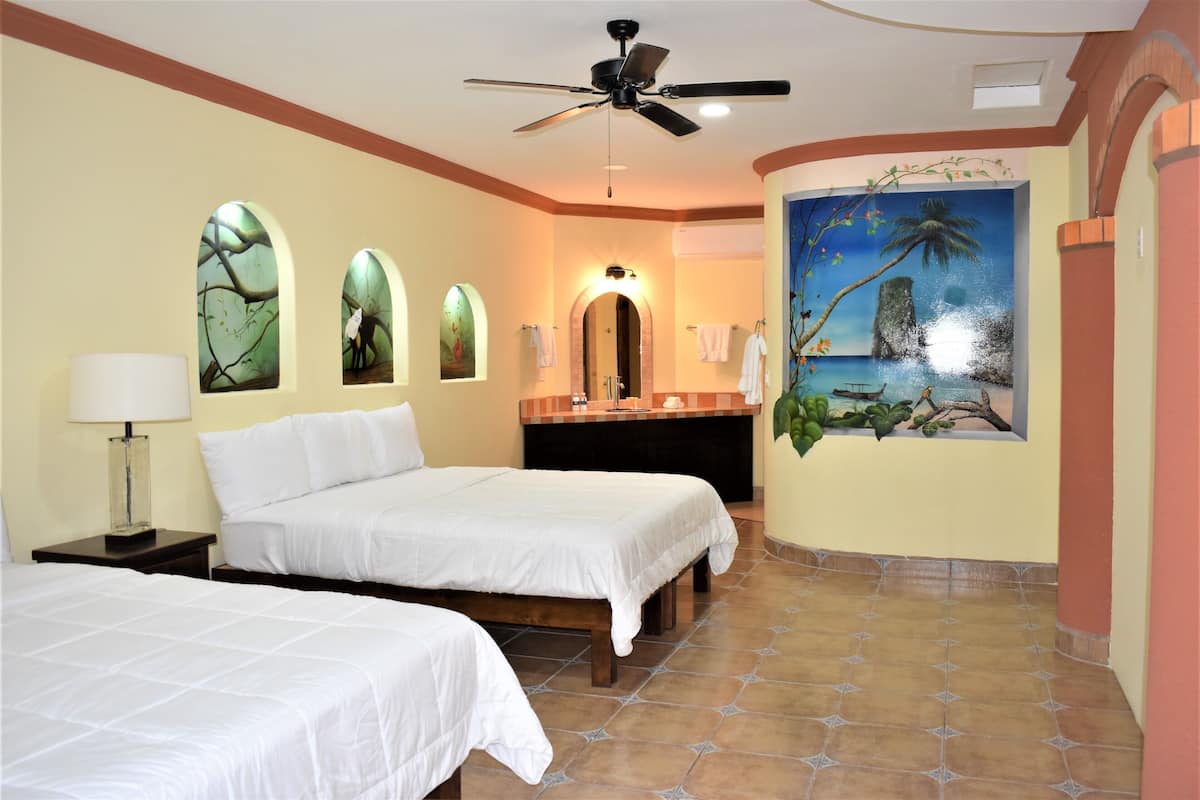 A hotel room with two beds, a ceiling fan, a painting of a tropical scene, and a tiled floor.