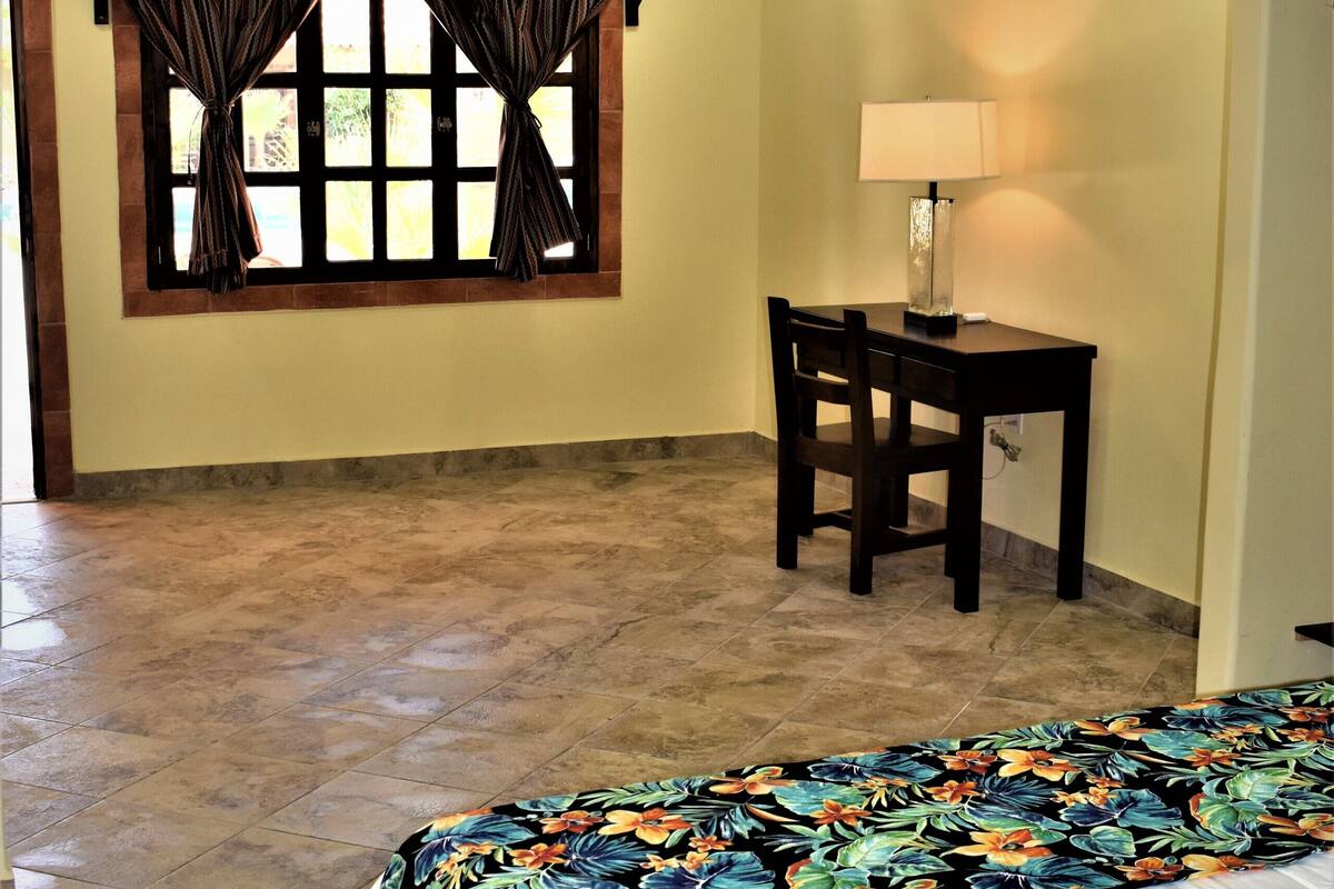 A bedroom with a bed, a desk, a chair, a lamp, a ceiling fan, and a window with curtains.