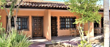 A building with a tiled roof, multiple doors, and windows. There are outdoor tables and chairs, and a palm tree.