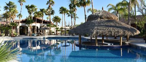 A swimming pool with a thatched-roof structure, surrounded by palm trees and a building with arches.