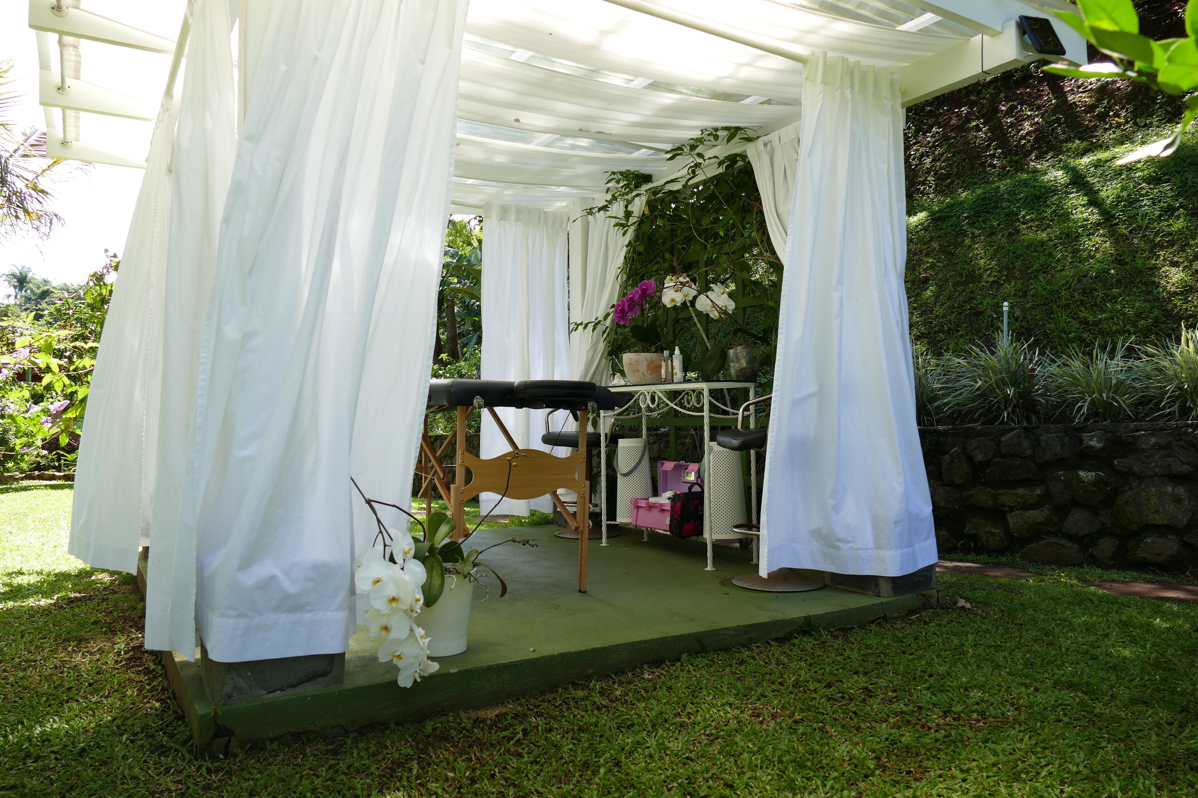 Een overdekt terras met een tafel en stoelen, omgeven door groen en een stenen muur.