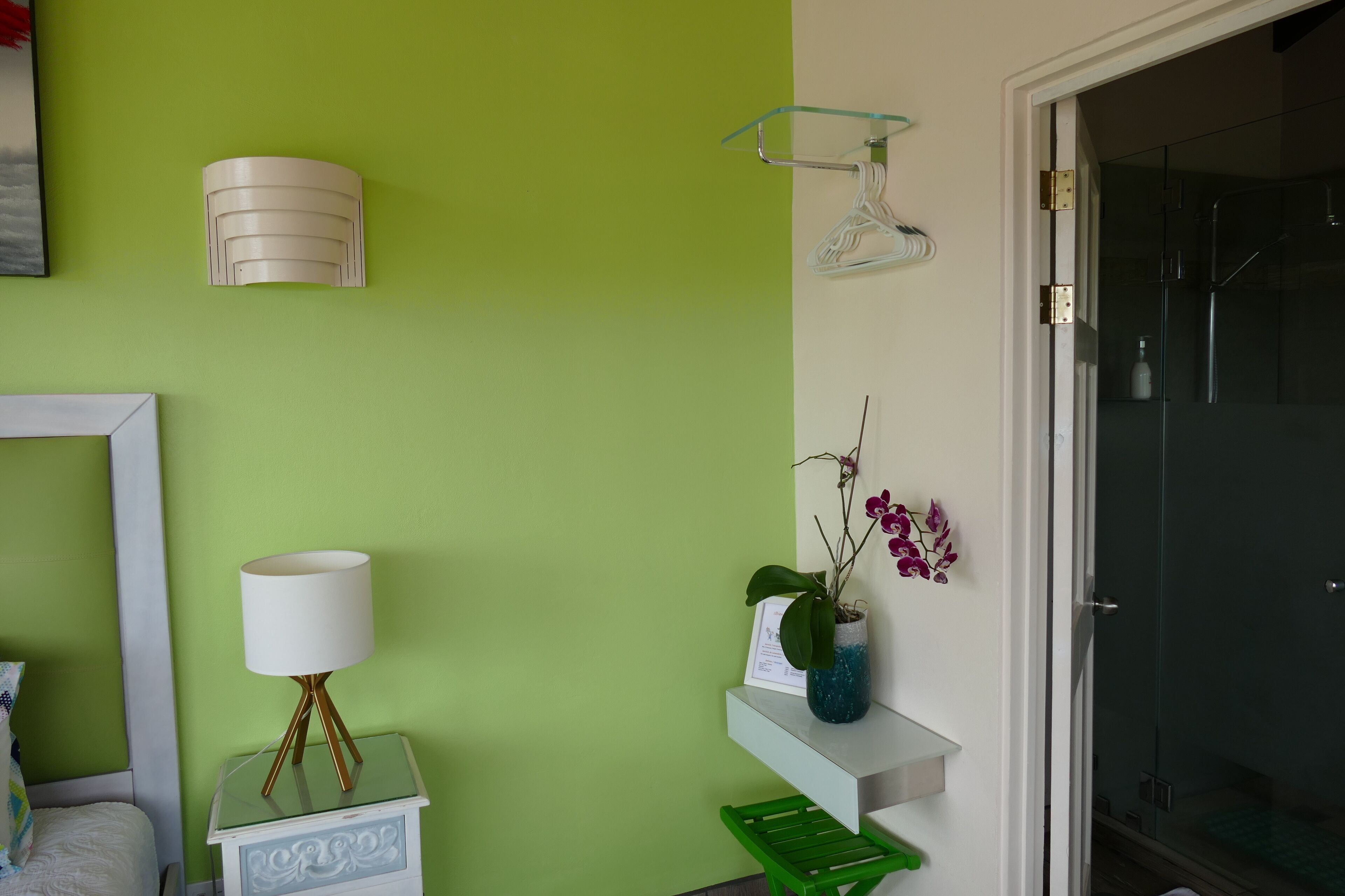 A room with a green wall, a white lamp, a small table with a lamp, a green chair, and a glass shower enclosure.