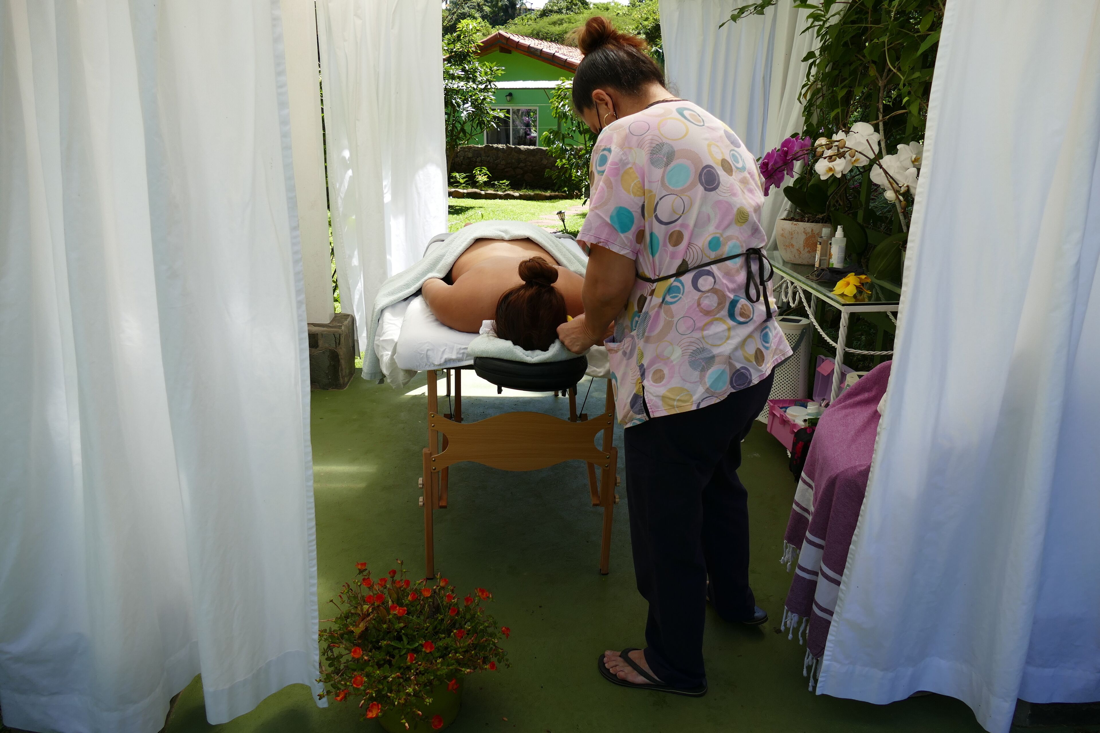 Una sesión de masoterapia en una habitación con piso verde, cortinas blancas y una silla de madera.