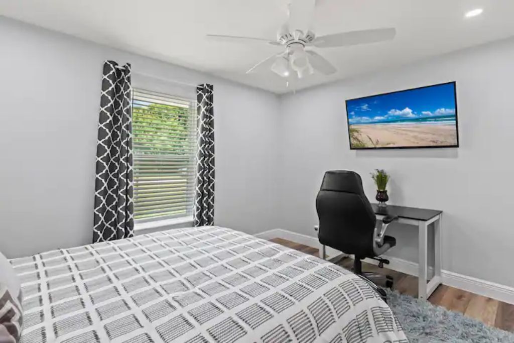 Ein Schlafzimmer mit Bett, Schreibtisch, Stuhl, Deckenventilator und einem Fenster mit Vorhängen.